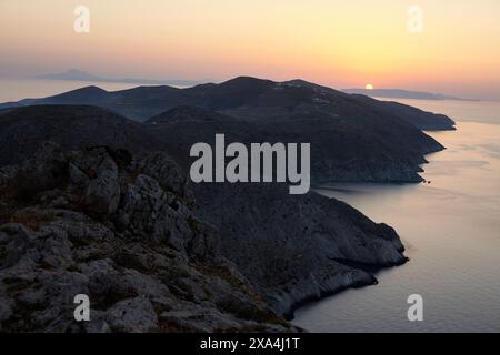 The sun sets behind a mountainous coastline, casting a warm glow over the serene seascape. Stock Photo