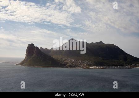 Coastal landscape featuring a seaside town nestled at the base of a dramatic mountain range under a partly cloudy sky. Stock Photo