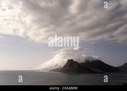 Clouds blanket the rugged peaks of a mountain beside a calm sea under a dramatic sky. Stock Photo