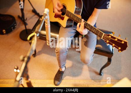 A musician is sitting on a stool playing an acoustic guitar in a warmly lit room with a microphone and another guitar in the background. Stock Photo