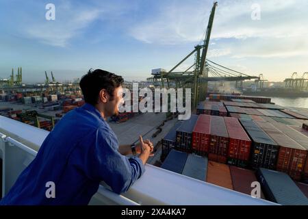 A man gazes out over a busy container port while taking notes, with shipping cranes and stacked containers in the background during dawn or dusk. Stock Photo