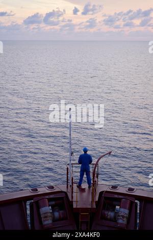 A person stands on the stern of a ship, gazing out at the vast ocean during twilight. Stock Photo