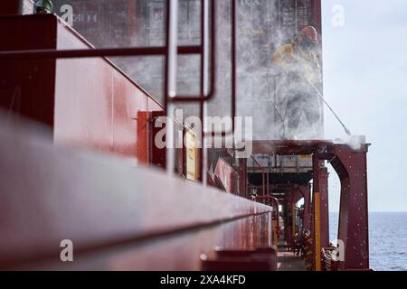 A worker in protective gear uses a high-pressure hose on the deck of a large ship, creating a mist of water around them. Stock Photo