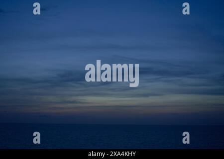 A calm ocean horizon under a twilight sky with subtle gradients from deep blue to orange near the horizon, and streaks of clouds above. Stock Photo