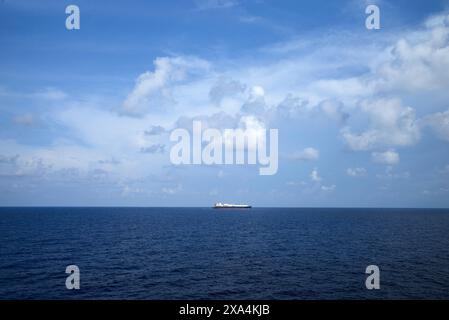 A cargo ship sits in the distance on a calm blue ocean under a vast sky scattered with white clouds. Stock Photo