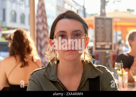 A young woman with glasses is looking directly at the camera with a serious expression, with blurred people and urban street life visible in the background. Stock Photo