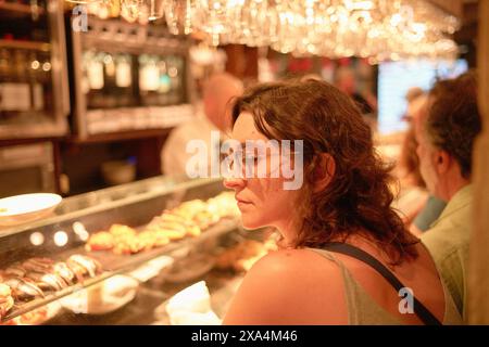 A woman is sitting at a bar with a thoughtful expression, gazing to her side, as the warm ambient lighting illuminates an array of tapas in the background. Stock Photo