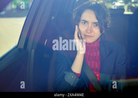 A woman in a red turtleneck rests her head on her hand while sitting in a car, looking through the window with a reflective expression. Stock Photo