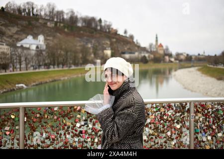 A person stands on a bridge over a river, smiling at the camera, with numerous love locks in the background. Stock Photo
