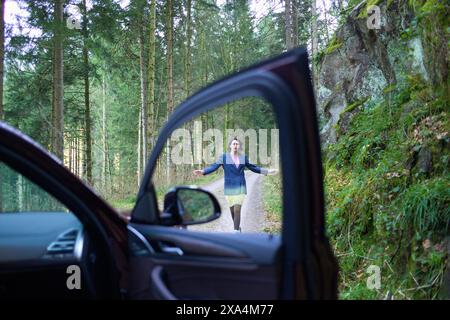A woman is standing in the middle of a forest road, viewed through an open car door from the driver's seat perspective. Stock Photo