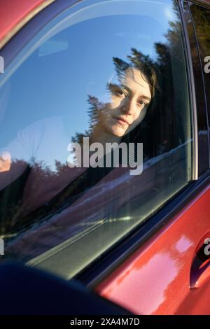 A person is looking through a car window, with their face partially obscured by reflections of trees. Stock Photo