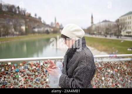 A person stands on a bridge overlooking a river with numerous love locks attached to the railing, while checking their smartphone. Stock Photo