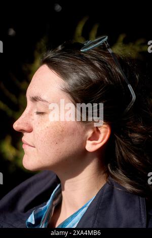 A woman with freckles, her eyes closed, and hair clasped with a blue clip, is basking in the sunlight with a serene expression. Stock Photo
