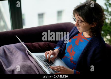 A person is sitting on a couch working on a laptop, dressed in a blue sweater with a red pattern, with a blanket over their legs. Stock Photo