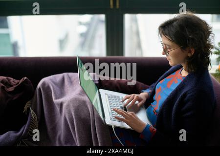 A woman sits comfortably on a purple couch with a blanket, focused on her laptop. Stock Photo