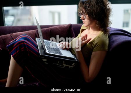A young woman with curly hair sits on a purple sofa, working on a laptop and holding an apple. Stock Photo