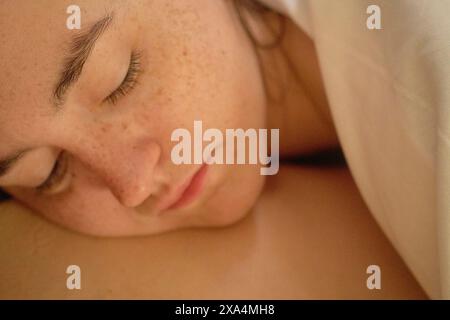 Close-up of a young woman sleeping peacefully with freckles on their face and eyes closed under warm lighting. Stock Photo