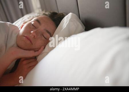 A young woman sleeping peacefully in a bed with white bedding and a subdued lighting environment. Stock Photo