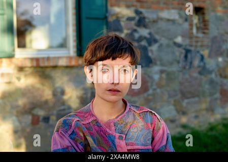 A portrait of a young woman with short hair and striking blue eyes wearing a patterned shirt, posed in front of a stone wall and window. Stock Photo