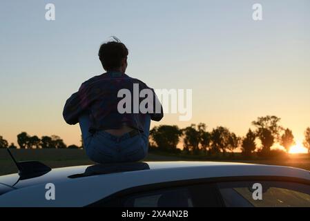 A young woman sits on the roof of a car, silhouetted against a sunset sky with trees on the horizon. Stock Photo