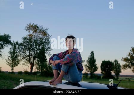 A young woman sits on the roof of a car during twilight, with trees in the background and a crescent moon visible in the sky. Stock Photo