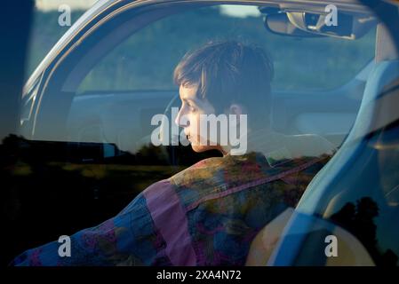 A young woman is sitting in the driver's seat of a car, with a thoughtful expression, looking out the window during sunset. Stock Photo