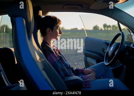 A young woman sits in the driver's seat of a car at sunset, gazing thoughtfully to the side with a calm expression. Stock Photo