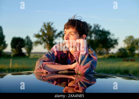 A young woman with short hair rests their chin on their hand while leaning on a reflective surface outdoors at sunset, displaying a contemplative expression. Stock Photo