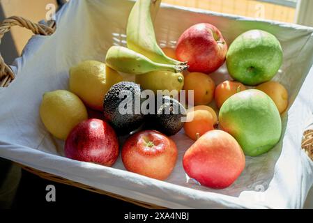 A variety of fresh fruits, including apples, lemons, a banana, avocado, and peaches, are neatly arranged in a woven basket by a window. Stock Photo