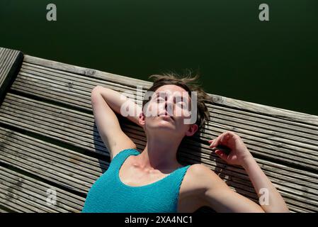 A young woman is sunbathing on a wooden dock over water, wearing a blue tank top with eyes closed and head resting back. Stock Photo