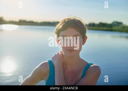 A woman with short hair in a blue dress stands by a body of water at sunset, with sunlight softly highlighting her hair. Stock Photo