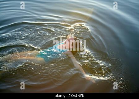 A young woman is swimming in calm water with sun rays reflecting on the surface. Stock Photo