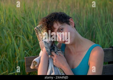 A young woman  with short hair sits outdoors resting their head on their hand, with tall grass in the background during sunset. Stock Photo