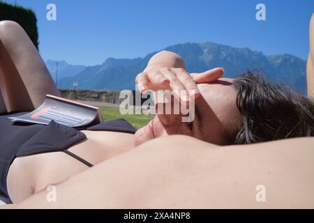 A person is lying down with their head resting on their arm, shielding their eyes from the sun, with mountains in the background. Stock Photo