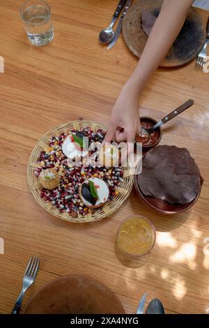 A person's hand reaching out to a colorful Mexican meal on a woven plate, accompanied by tortillas, sauces, and a drink on a wooden table. Stock Photo