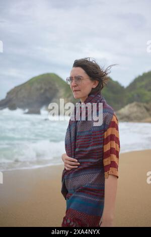 A woman stands on a sandy beach with her hair blowing in the wind, facing the sea with a rocky outcrop in the background. Stock Photo
