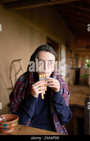 A young woman is sitting at a rustic wooden table playing a wooden pan flute. They are wearing eyeglasses, a plaid shawl, and have a contemplative expression. Stock Photo