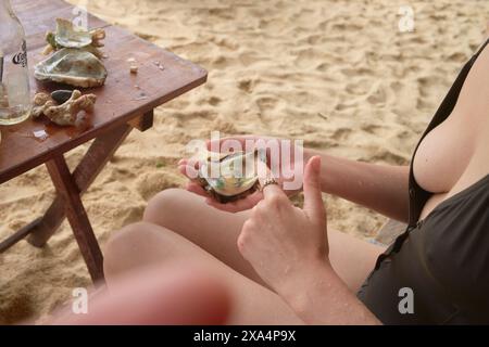 A person is sitting at a wooden table on the beach, holding a colorful glass ornament in their hands with seashells scattered on the table surface. Stock Photo