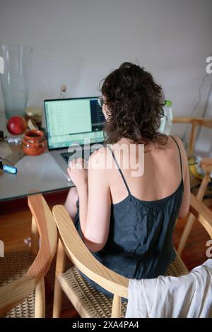 A young woman with curly hair is sitting at a wooden dining table and working on a laptop, with a smartphone and fruits nearby. Stock Photo