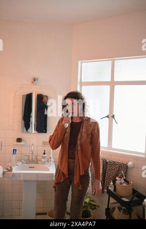 A young woman stands in a well-lit bathroom, brushing their teeth and looking at the camera with a contemplative expression. Stock Photo