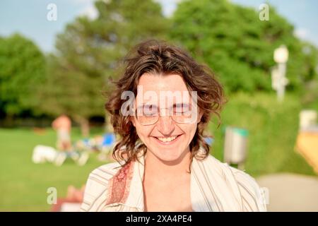 A smiling young woman with curly hair and glasses is standing outdoors on a sunny day, with blurred greenery and people in the background. Stock Photo