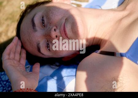 A young woman is lying on their back outdoors, smiling slightly at the camera with sunlit grass in the background. Stock Photo