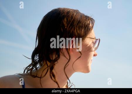 Side profile of a young woman wearing sunglasses with wet hair against a clear blue sky. Stock Photo