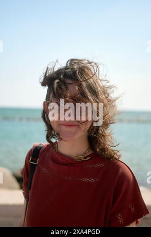 A woman is standing outdoors with the sea in the background, her hair tousled by the wind, wearing glasses and a red top with a subtle smile. Stock Photo