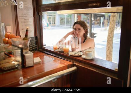 A woman is sitting at a bar table beside an open window, looking pensively outside with drinks and snacks in front of her. Stock Photo