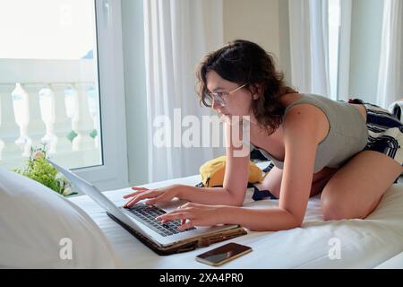 A young woman is reclining on a bed while using a laptop, with curtains and a balcony railing visible in the background. Stock Photo