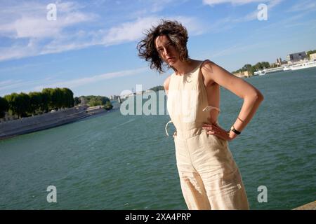 A person stands confidently by a riverside with hair blowing in the wind, wearing a light-colored jumpsuit against a blue sky. Stock Photo