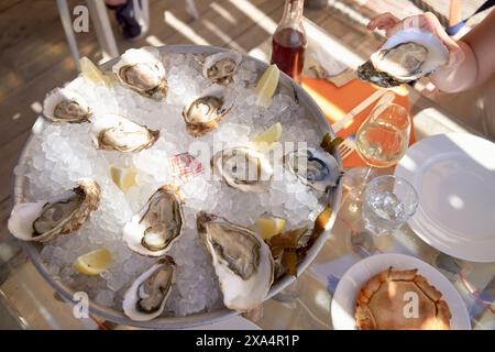 A platter of fresh oysters on ice is served on an outdoor table alongside a glass of white wine, a bottle, and an empty plate. Stock Photo
