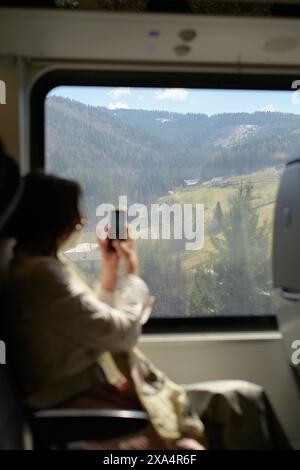 A person is seated inside a train, capturing the scenic mountainous landscape outside the window with their smartphone. Stock Photo