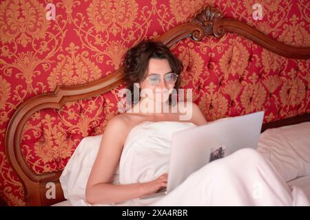 Smiling woman with glasses using a laptop in bed with red ornate wallpaper in the background. Stock Photo